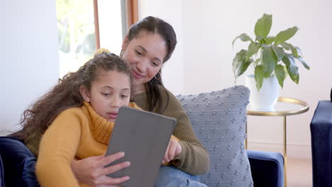 Happy-biracial-mother-and-daughter-embracing-on-sofa-and-using-tablet-in-sunny-living-room