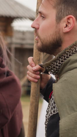 bearded knight and young woman with flute stand against white tent in courtyard. couple stands near wooden arch looking at territory on blurred background closeup