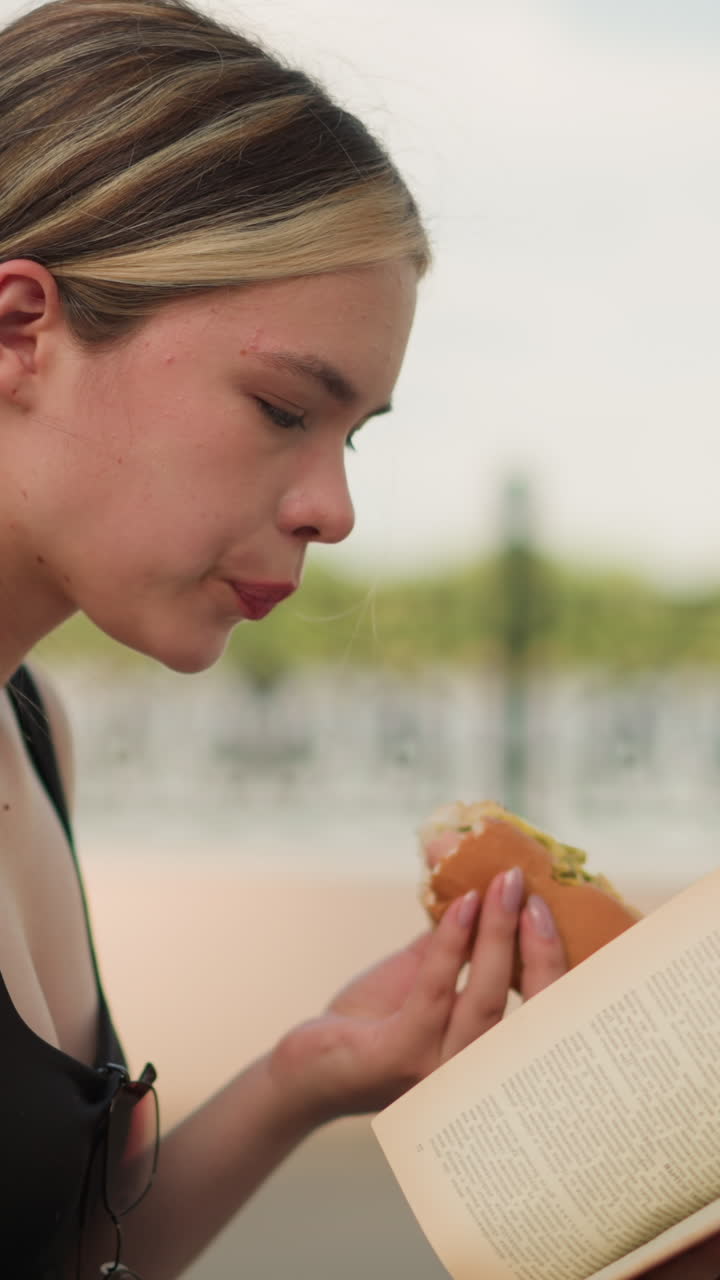mujer con camiseta negra sentada al aire libre, disfrutando de un bocadillo mientras lee un libro, con una valla decorativa y un cielo brillante en el fondo, combinando casualmente la lectura y la comida