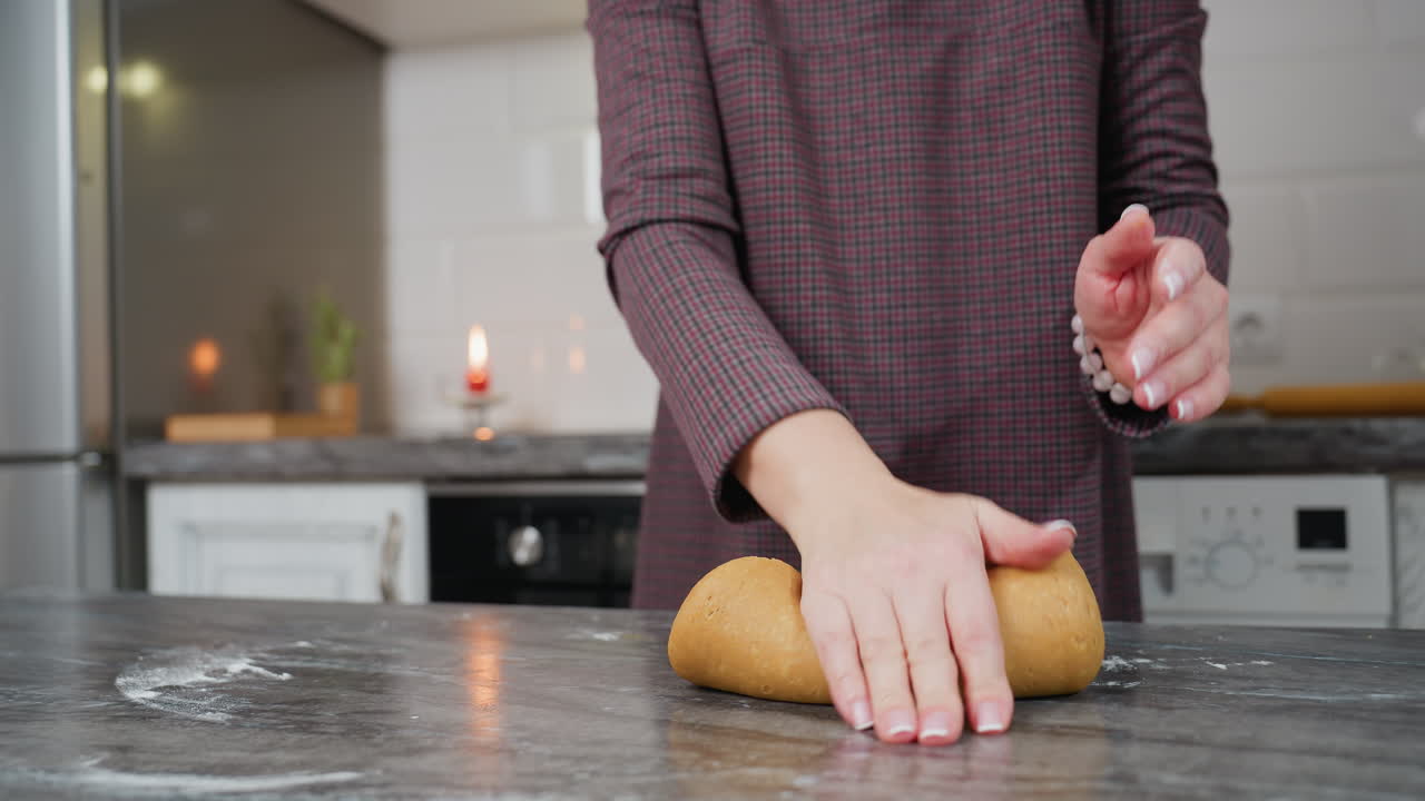 Woman with bracelet on left hand kneads fresh dough on kitchen counter, preparing homemade baked goods, modern kitchen setting features rolling pin, red candle, white cabinets, and warm lighting