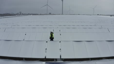 technician inspects snow-covered solar panels at a renewable energy farm with wind turbines
