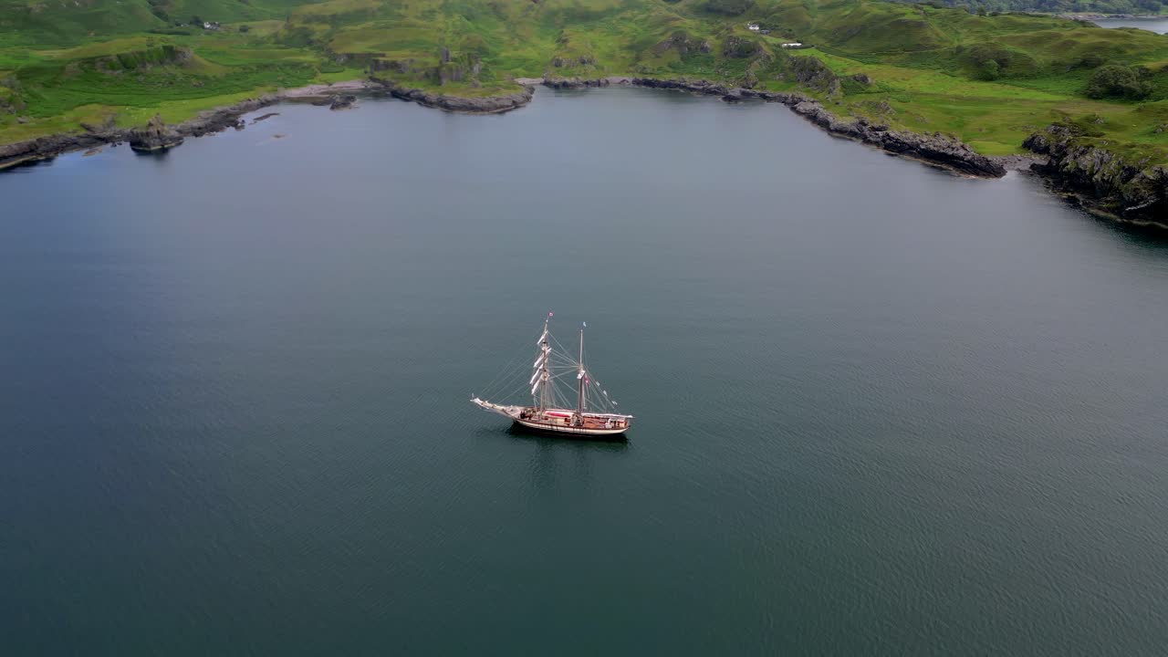 Tampilan depan udara kapal layar tinggi bersejarah berlayar di sepanjang garis pantai Skotlandia yang indah. Kapal layar tradisional dengan layar putih di dekat pantai berbatu hijau. Pemandangan maritim sinematik di perairan Hebrides Skotlandia.