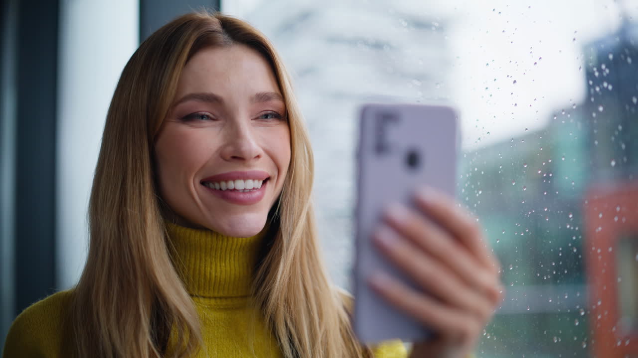 Blonde watching cellphone social media standing by apartment window closeup