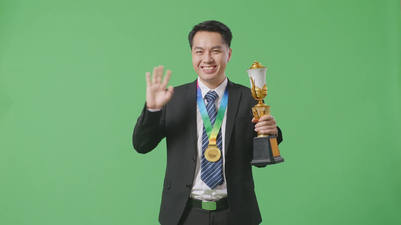 슈트와 타이를 입은 아시아 사업가, 금메달과 트로피를 들고 손을 흔들며 카메라에 미소를 지으며 스튜디오의 녹색 스크린 배경에서 첫 번째 우승자로