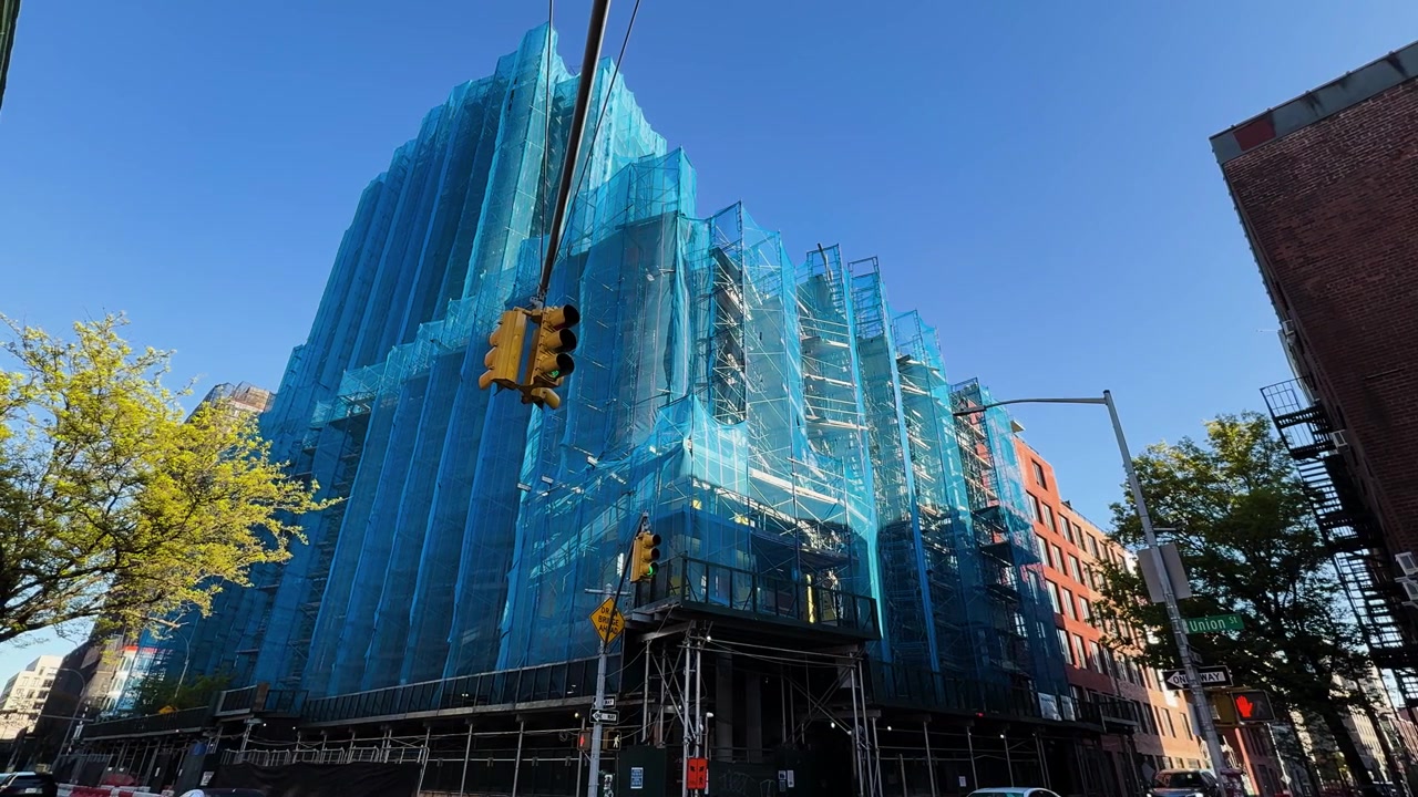 Il traffico scorre davanti a un cantiere edile con un edificio coperto da una rete di sicurezza blu a maglie in una giornata di sole.