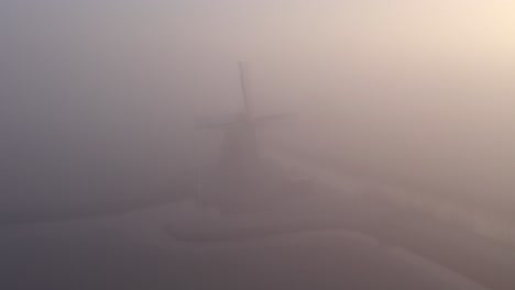 Flying-backwards-away-from-Cornwerder-molen-windmill-during-a-foggy-morning,-aerial