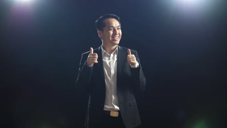 smiling asian speaker man in business suit showing thumbs up gesture and looking around while standing in the black screen studio