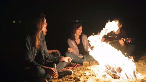 Young-cheerful-people-sitting-by-the-bonfire-late-at-night,-playing-guitar,-singing-songs-and-drinking-beer.-Cheerful-friends-talking-and-having-fun