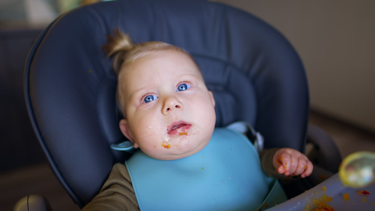 Baby with plump cheeks and blond hair sits in the chair. Portrait of a child being fed from spoon. Close up.