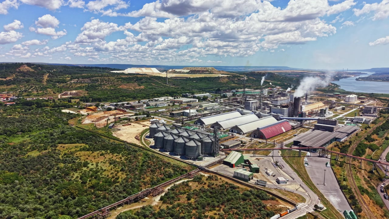 Factory located near a river surrounded by hills and fields with clouds in the blue sky and a view of large storage silos and industrial buildings in the area