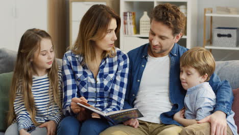 cheerful family of parents and little son and daughter sitting all together on the couch and flipping the pages of the english book