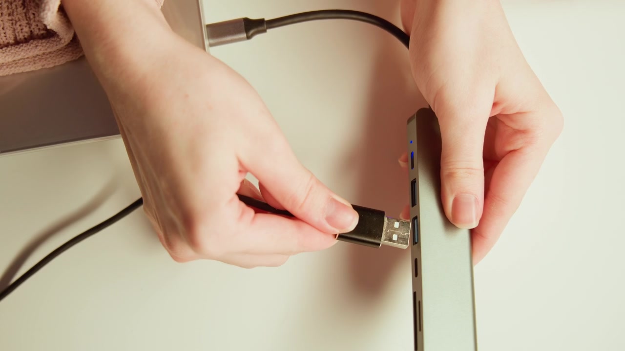 Top view close-up shows hands guiding USB connector into metallic hub across beige tabletop, with black braided cable, warm overhead light, and subtle motion blur during careful alignment and