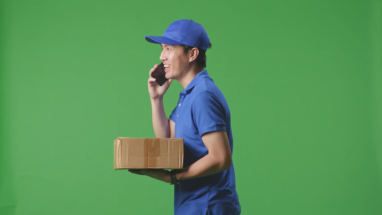 Side View Of Asian Male Courier In Blue Uniform Walking And Talking On Smartphone While Delivering A Carton On Green Screen Background In The Studio