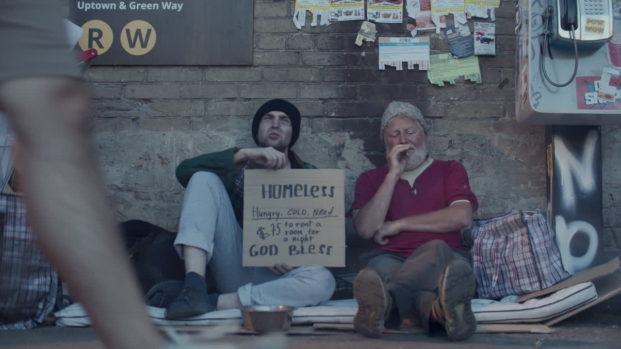 Two Homeless Men Sitting with Cardboard Sign on Street, Talking and Smoking