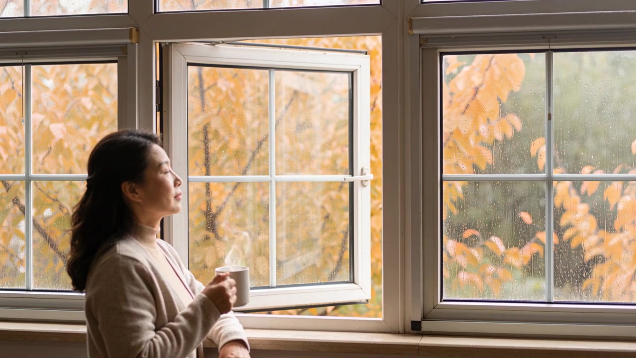 Standing woman in tan cardigan holding mug watching rain falling on window orange leaves copy space