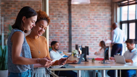 Two-Female-Business-Colleagues-With-Digital-Tablet-And-Documents-Meeting-In-Busy-Office