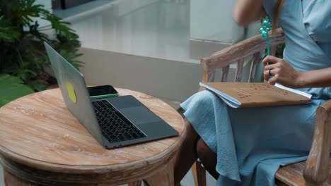 a girl in a blue dress remotely online working behind a laptop and looking into the screen the backyard with green plants