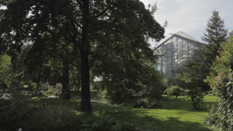 Lush-greenery-with-a-modern-glasshouse-in-a-serene-park-setting,-sun-peeking-through-trees