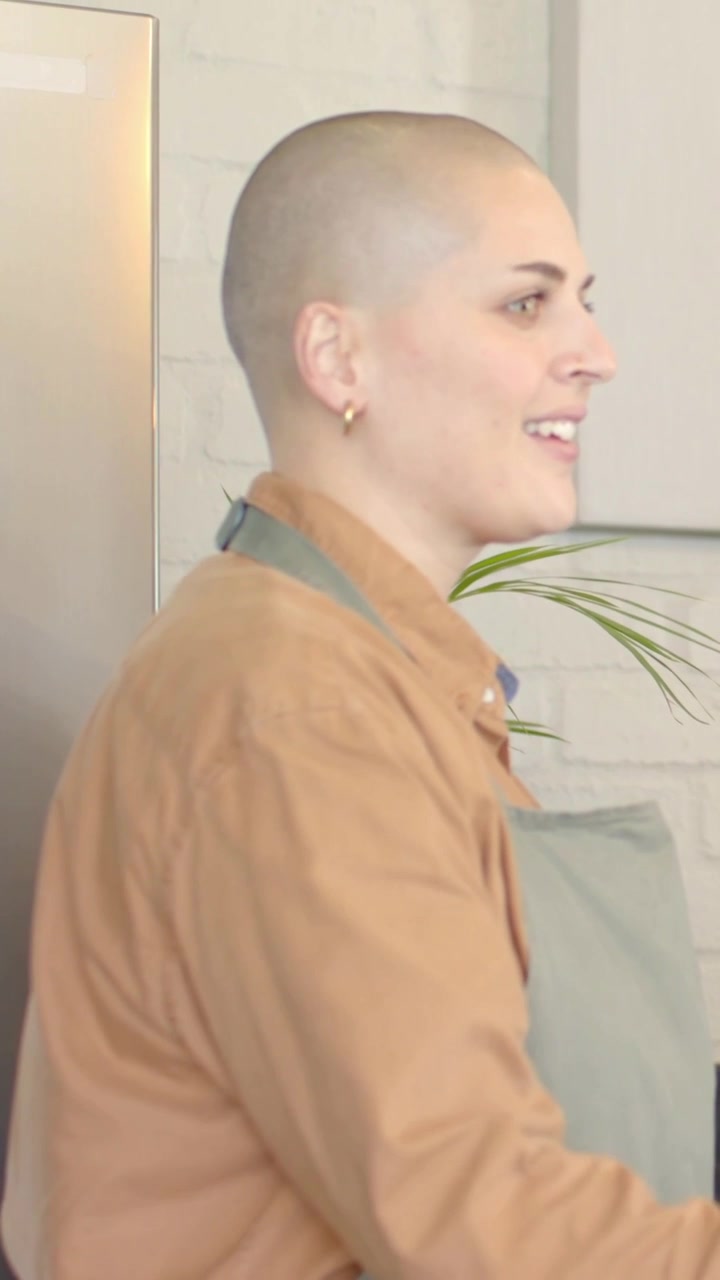 Video verticale: Donna sorridente con grembiule verde che parla in cucina, reagendo a una mano blu fuori campo.