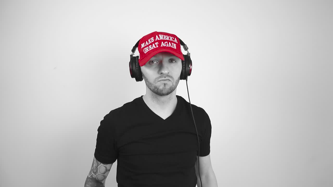 Grayscale footage of a tattooed man with a red MAGA Make America Great Again Trump hat putting on large studio earphones to listen to music and enjoys the music while moving around slowly.