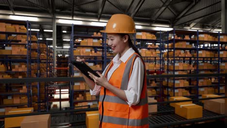 vista lateral de una ingeniera asiática con casco de seguridad de pie en el almacén con estantes llenos de bienes de entrega. tomando nota en la tableta en el almacenamiento