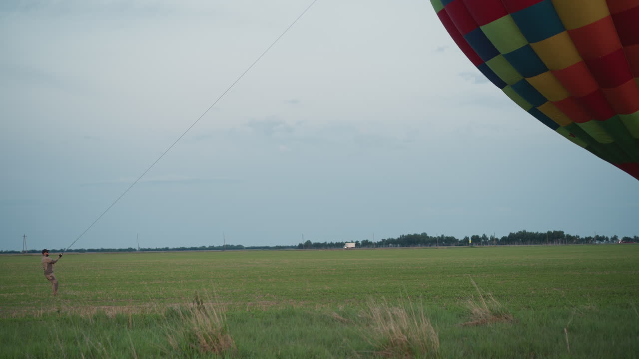 man gripping rope to tether vibrant hot air balloon on wind-swept green field while distant vehicle crosses horizon under pale sky capturing dynamic contrast between human effort