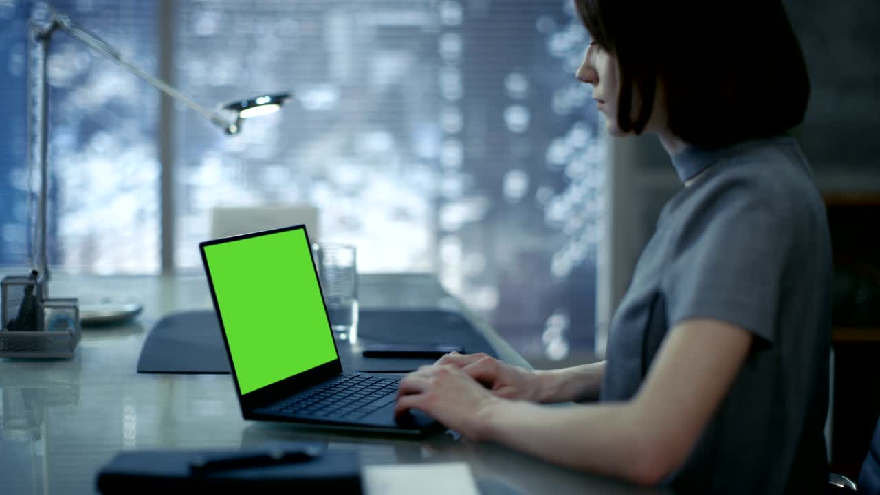 hermosa, elegante joven mujer de negocios se sienta en su mesa de trabajo y escribe en una computadora portátil con pantalla de maqueta verde. su oficina se ve moderno con connotaciones oscuras, gran ciudad detrás de la ventana.