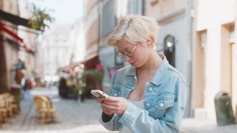 mujer rubia sonriente usando teléfono inteligente escribiendo mensajes de texto desplazando aplicación de redes sociales en la calle de la ciudad