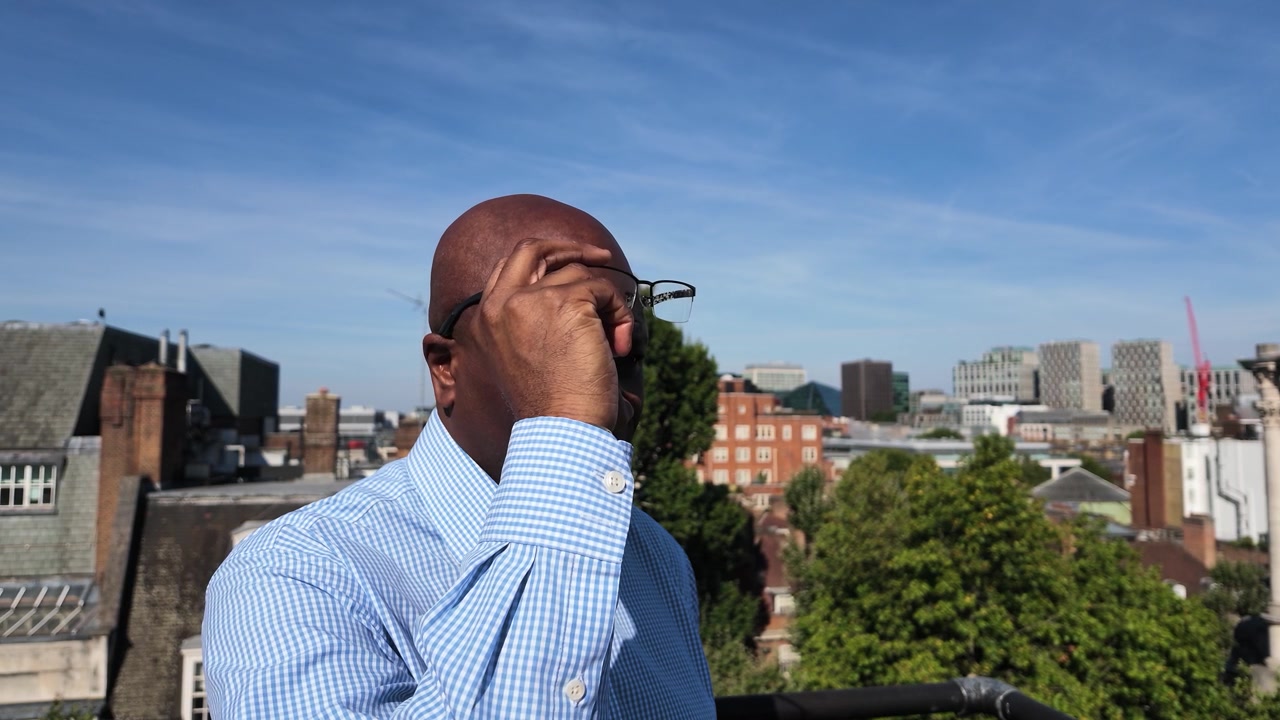 Male employee on a city rooftop slowly takes off his glasses in bright midday light, with buildings and trees in the background.