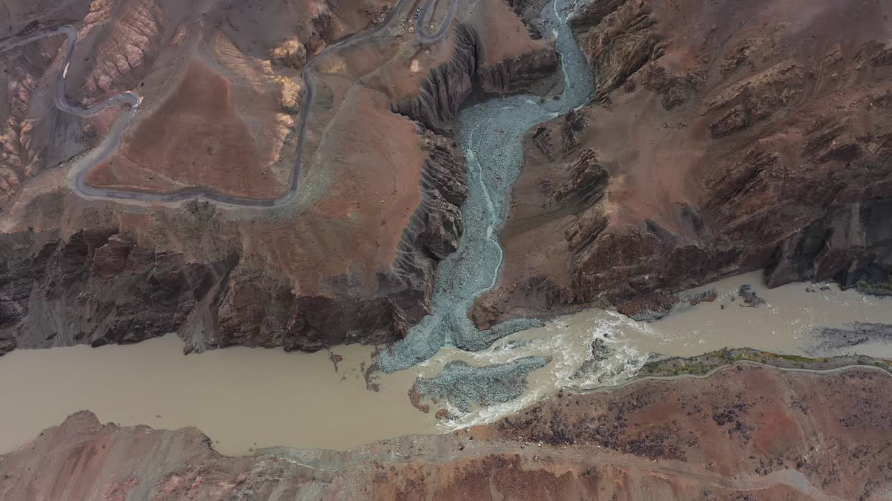 La gola del fiume Indo nel Ladakh attraversa un terreno montuoso arido, rivelando modelli di sedimenti e strade tortuose. Un movimento fluido del drone guarda in basso al punto di confluenza glaciale da alta quota.