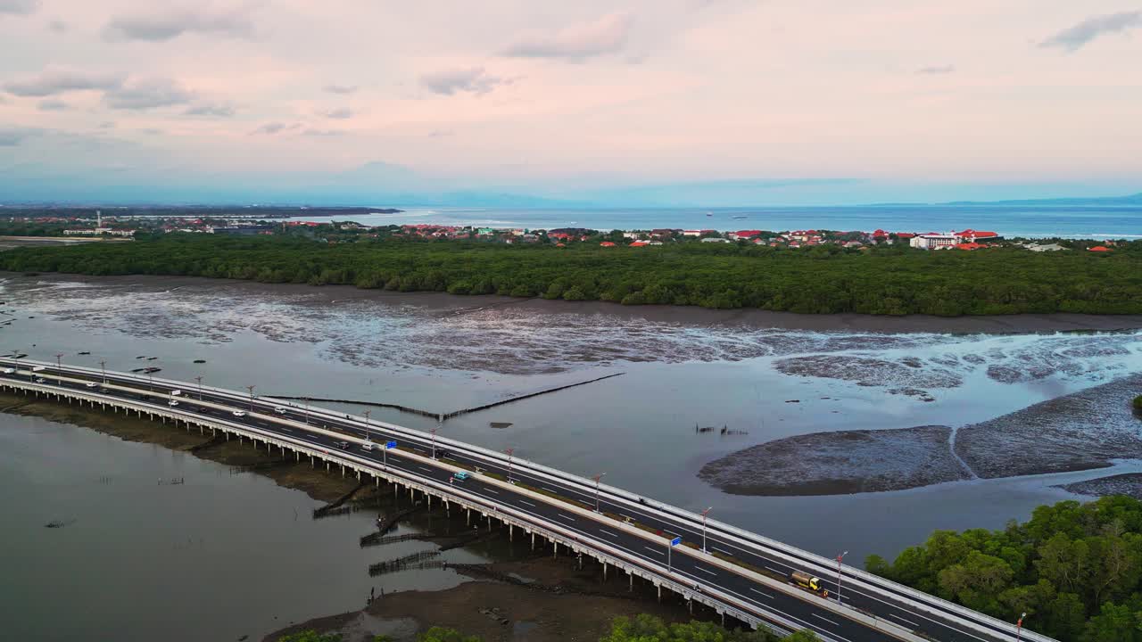 Aerial drone footage captures the Bali Mandara Toll road bridge extending across wide muddy flats revealed at low tide with a tropical Indonesian landscape creating a striking coastal highway scene