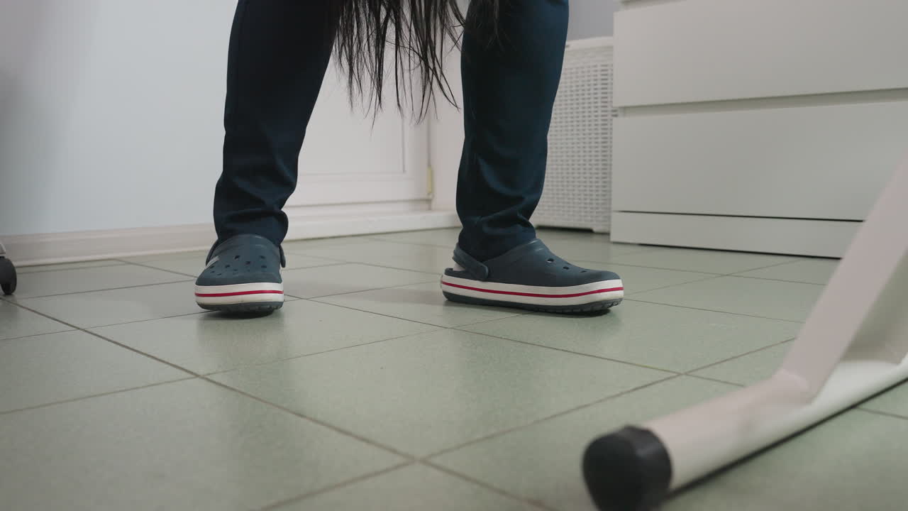 Beautician wearing clinical crocs moves beside spa bed as long dark hair strands fall from client onto tiled floor in bright treatment room showing storage cabinet and white basket in background
