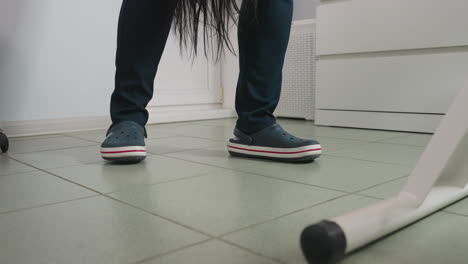 beautician wearing clinical crocs moves beside spa bed as long dark hair strands fall from client onto tiled floor in bright treatment room showing storage cabinet and white basket in background