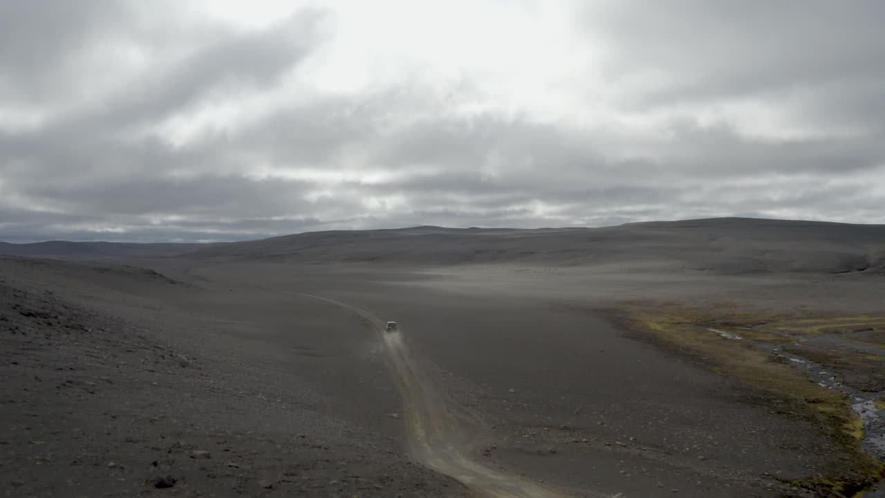 Aerial view of a vehicle kicking up dust while traversing a sprawling black volcanic sand desert with distant mountains in Iceland