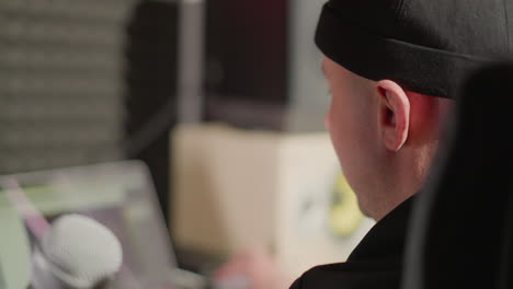 close-up side shot of a man wearing a black cap, with his ear prominently visible. a microphone is in the blurred background, suggesting a recording or audio production setting