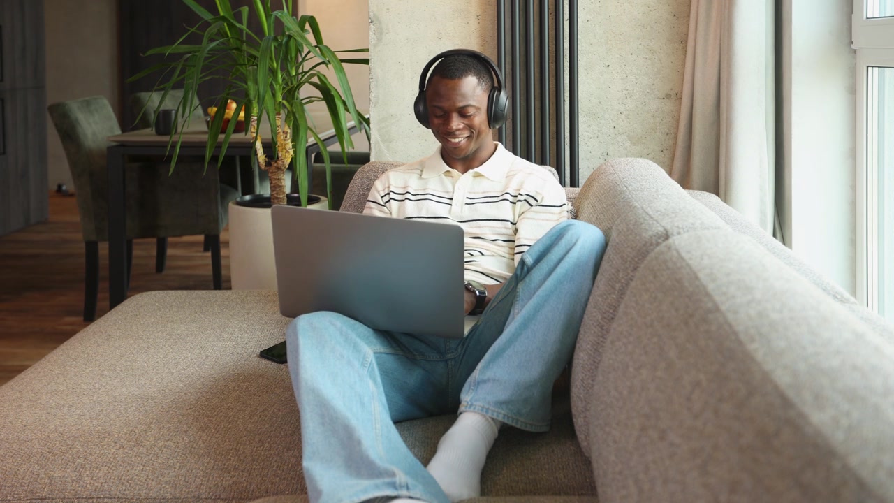 Young Black Man in Headphones Using Laptop on Couch at Home