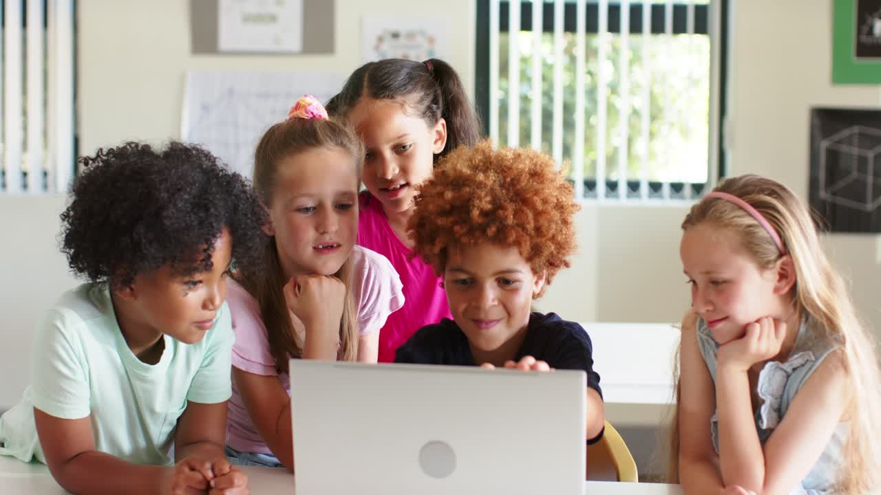 Verschiedene Kinder in T-Shirts beugen sich im Klassenzimmer um einen Laptop, reagieren auf Inhalte, während eines tippt.