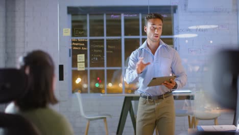 Biracial-businessman-using-tablet-and-glass-wall-presenting-to-colleague-at-night-in-office