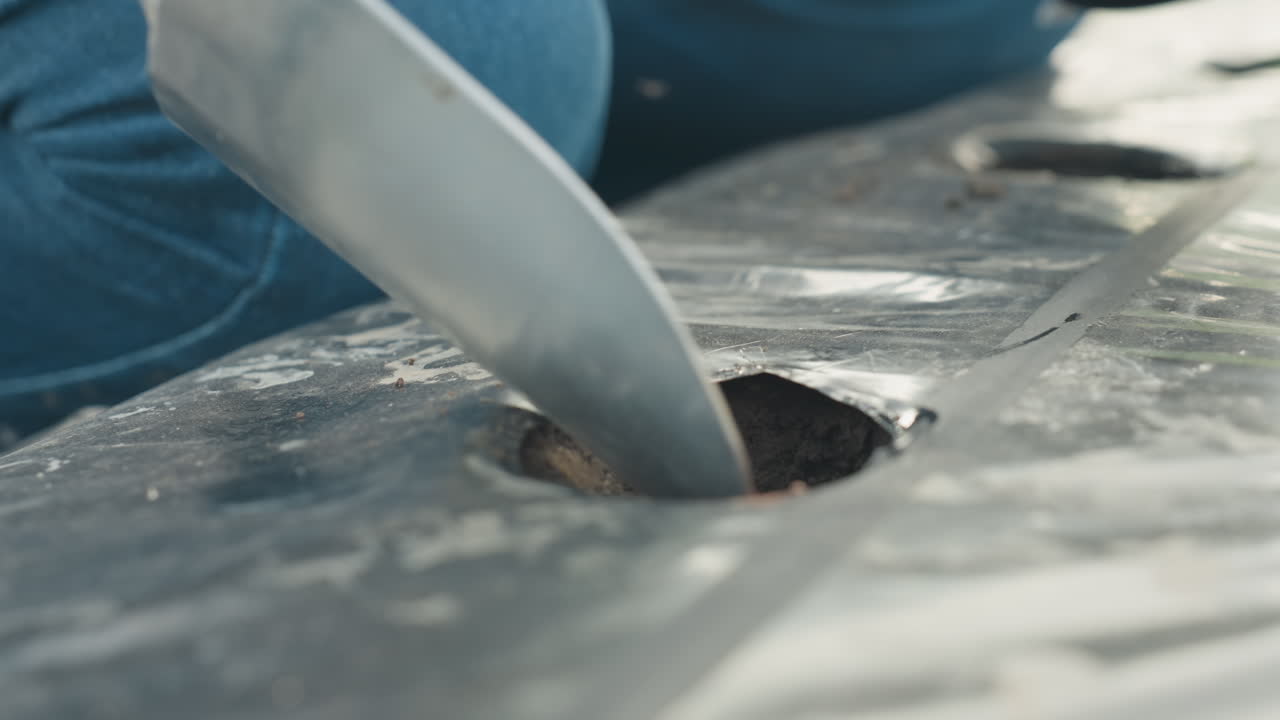 close up gloved hand drops seeds with shovel into soil through plastic mulch creating precise trench for planting young crops under dewy morning light in outdoor farm bed full of growth and nurture