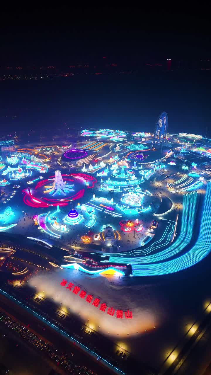 Vertical drone night shot of Harbin Ice Festival 2025, showcasing vibrant illuminated ice structures, colorful light displays, and a lively winter spectacle. China