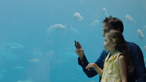 father-with-little-girl-at-aquarium-taking-photos-of-fish-using-smartphone-enjoying-marine-life-swimming-in-tank-having-fun-at-oceanarium-sharing-on-social-media