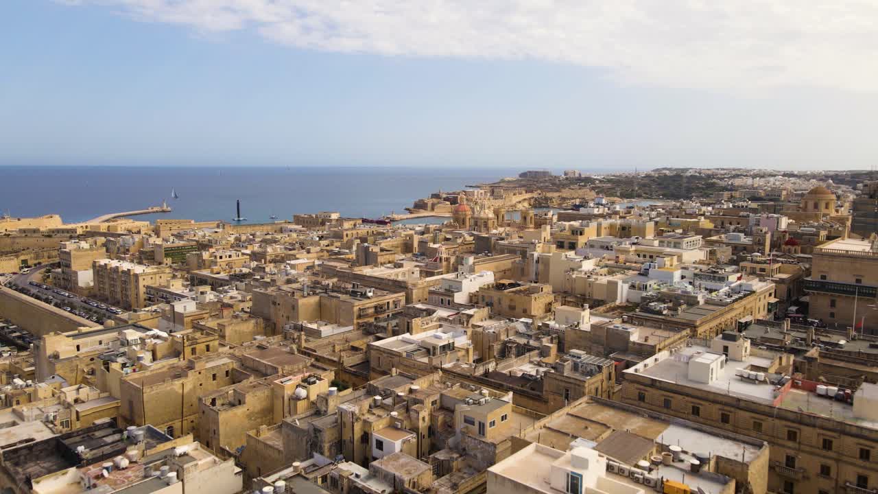 Epica prospettiva aerea con drone oltre la cupola barocca dorata e la croce prominente di una chiesa di La Valletta verso la costa mediterranea, la penisola del forte storico e l'orizzonte del mare blu profondo