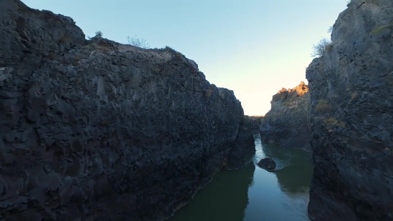 Dynamisk FPV-dronevideo som svever gjennom en dramatisk elvekløft i La Payunia-reservatet, Mendoza. Robuste vulkanske klipper, slyngete vannløp, lys i den gylne timen. Luftopptak av uberørt natur.