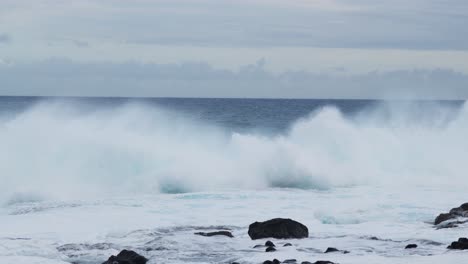 a turquoise wall of surf gathers and pitches toward a rocky shoreline, wind shearing spray from the crest as foamy white water surges across the frame with raw, restless energy