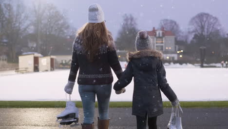 mamá de mediana edad y su hija de 8 años caminan de la mano en un día de invierno nevado a la pista de patinaje sobre hielo para practicar patinaje sobre hielo para actividades de unión como familia durante la diversión navideña