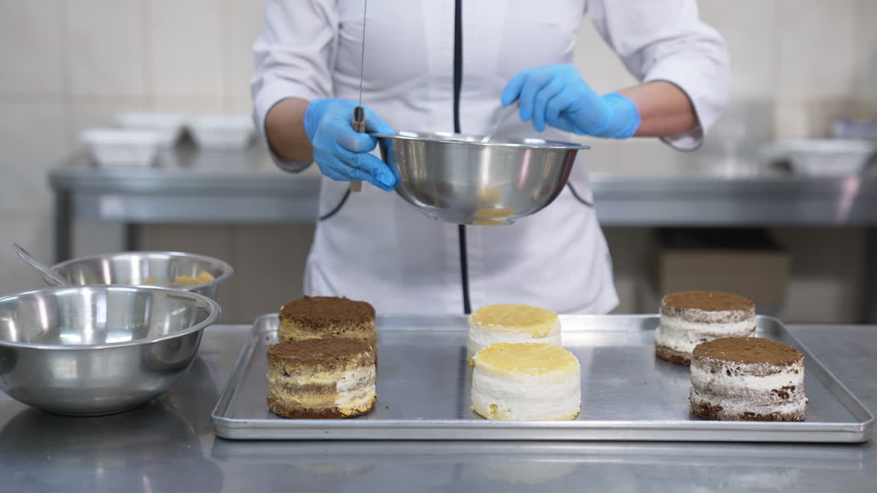 Little round cakes are put on the metal tray. Confectioner puts spoonfuls of cream on top of the cakes. Close up.