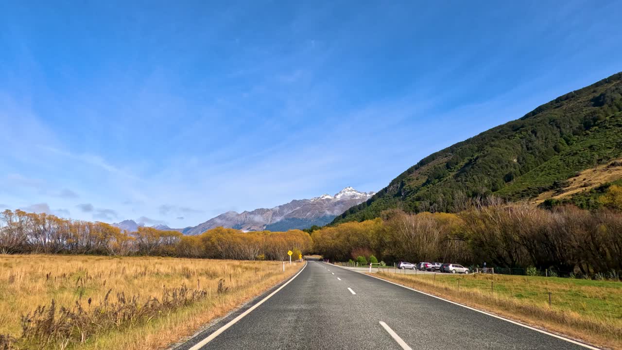 Vehicle travels along rural Routeburn road, passing golden autumn trees under bright daylight, wide angle