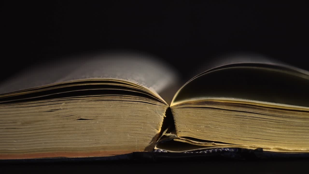 Slow motion, close up of an old, vintage, open book with pages turning on the wind, on a dark room with few light, with black surroundings