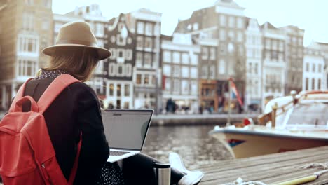 4k 30s happy businesswoman working with laptop. amsterdam. lady with red backpack sits on beautiful river embankment