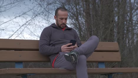 Closer-shot-of-young-bearded-man-with-short-hair-sitting-on-a-park-bench-on-a-cloudy-fall-day-and-playing-with-his-smart-phone-in-slow-motion